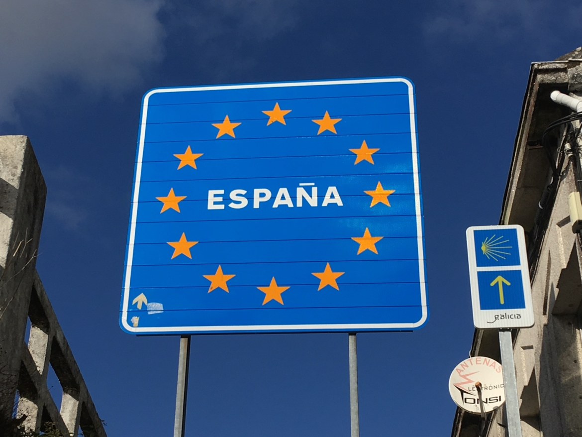 The blue España border sign beside a camino waymarker for Galicia