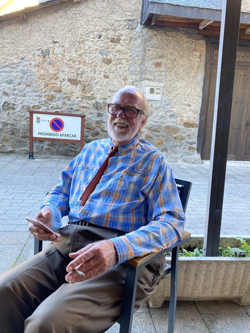 An older pilgrim with a white beard smiling over a cigar at a café