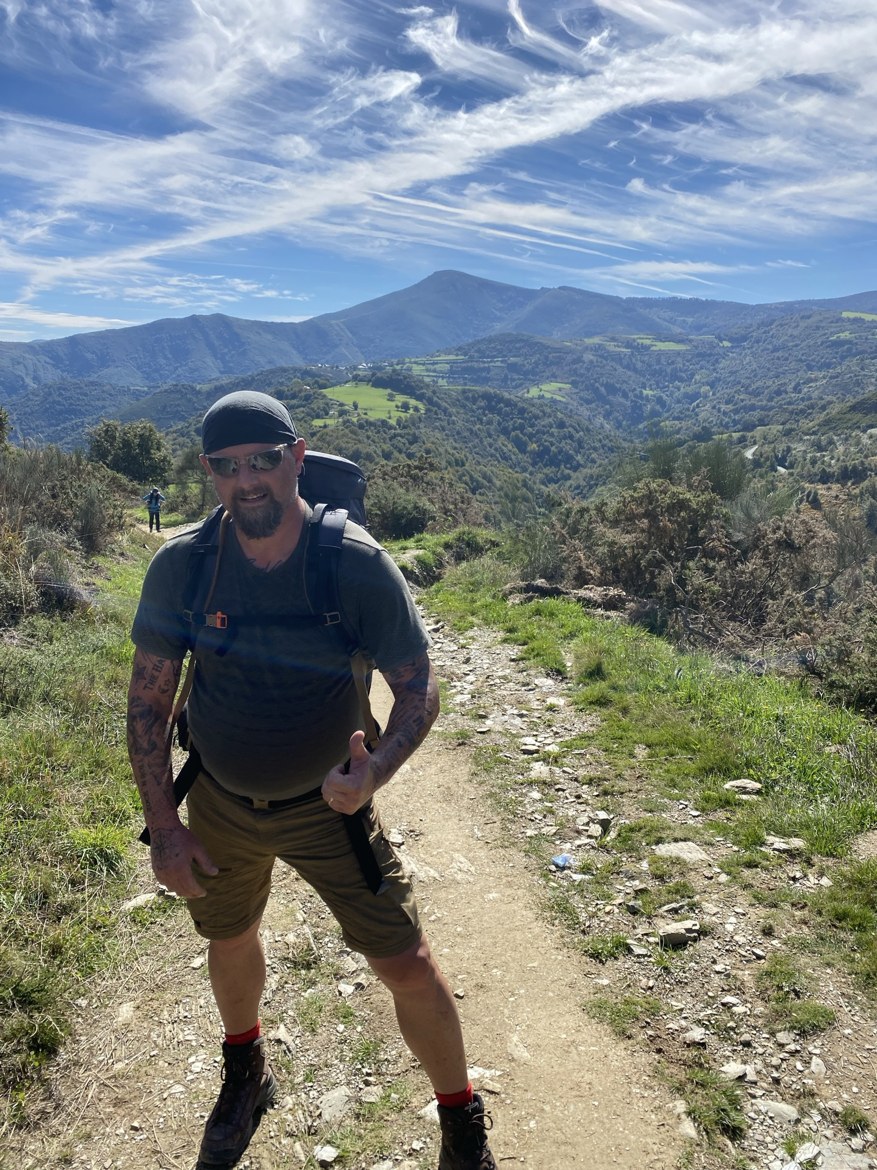 A pilgrim giving a thumbs up on a mountain trail above green valleys