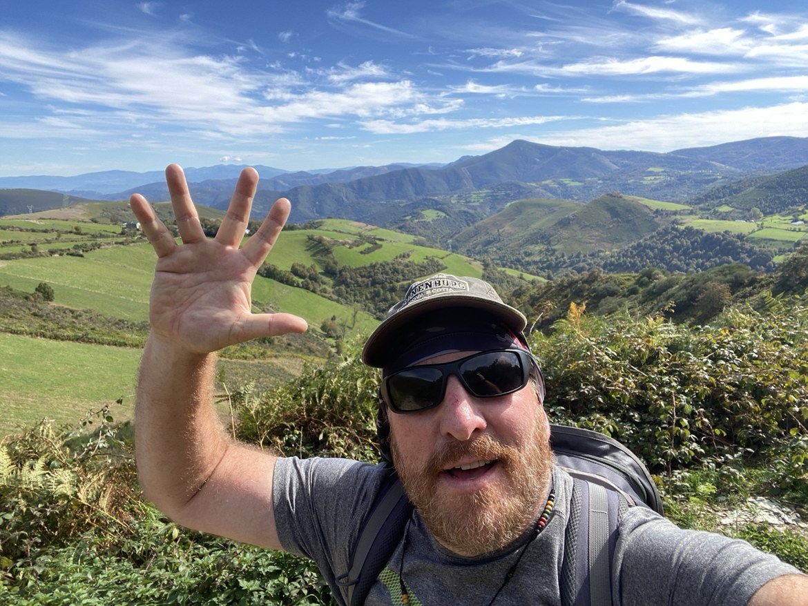 A pilgrim waving at the camera on a mountain trail above green valleys