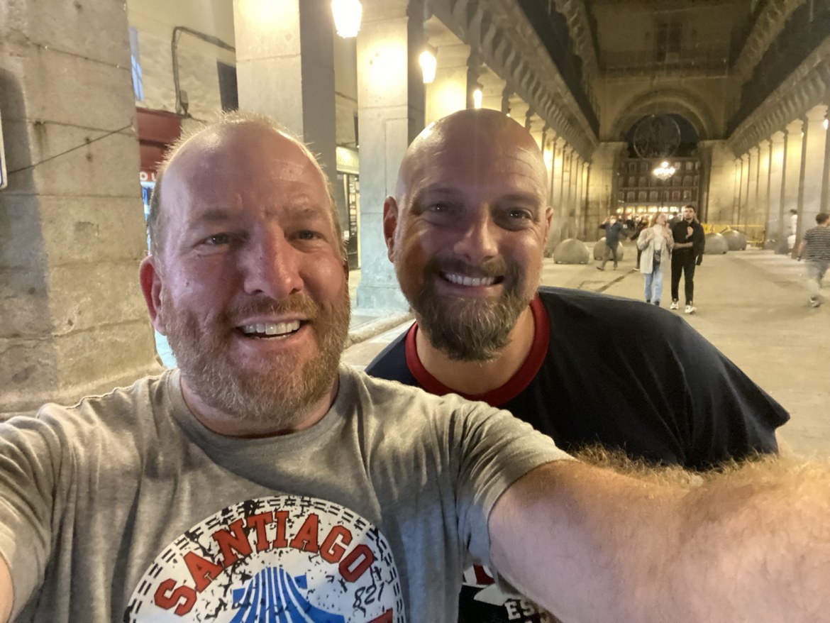 Two pilgrims taking a selfie in a grand arched plaza at night