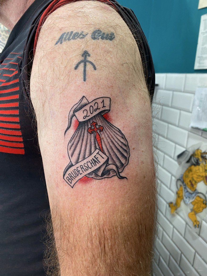 A fresh tattoo of a scallop shell with a red cross, reading 2021 Bruderschaft