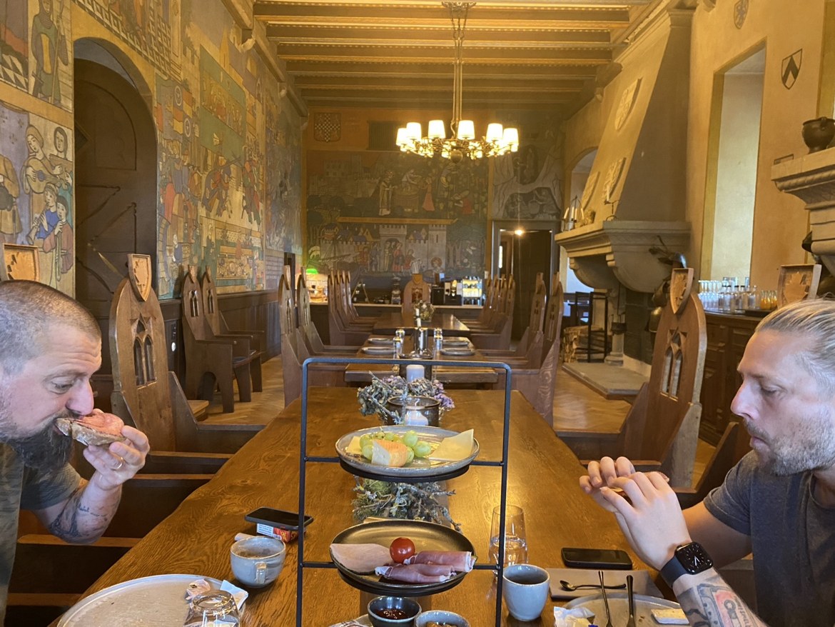 Nicki and Dennis eating breakfast at a long table in a mural-painted castle hall