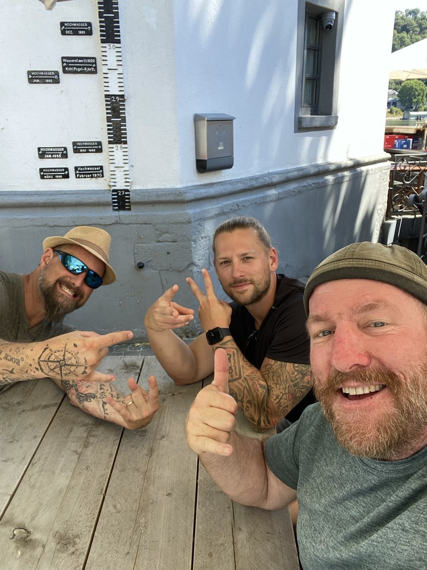 The three brothers at a table beneath historic flood-level markers on a wall in Koblenz