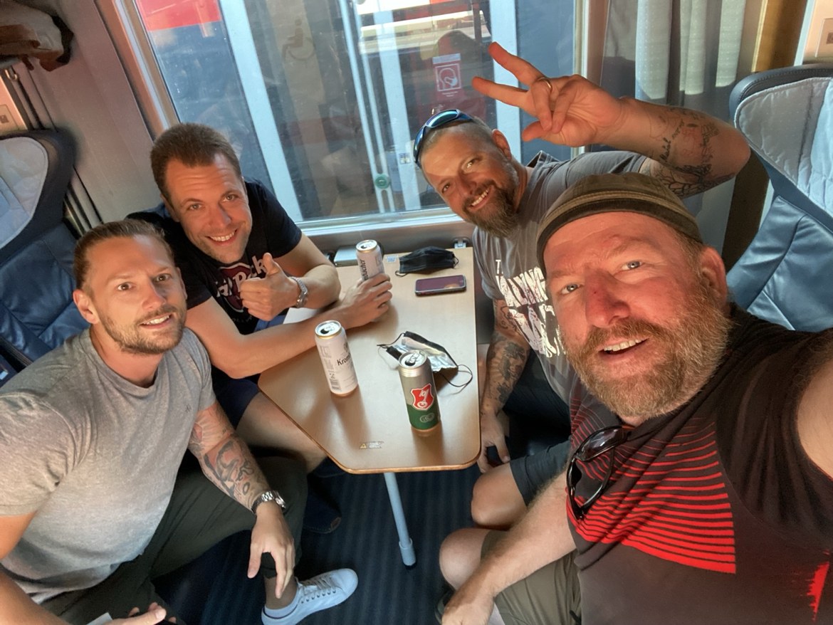 Four friends around a train table with cans of Krombacher