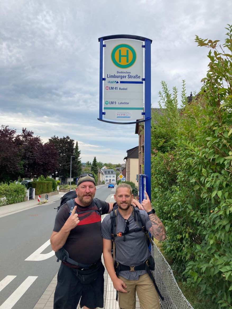Mike and Dennis with backpacks under a bus-stop sign on Limburger Strasse in Dietkirchen