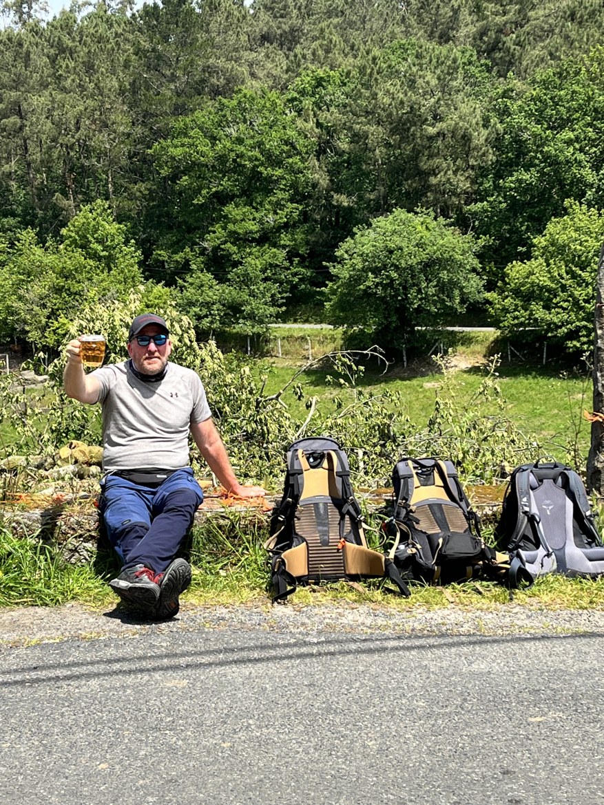 Mike sitting on a wall raising a glass of beer, the three backpacks lined up beside him