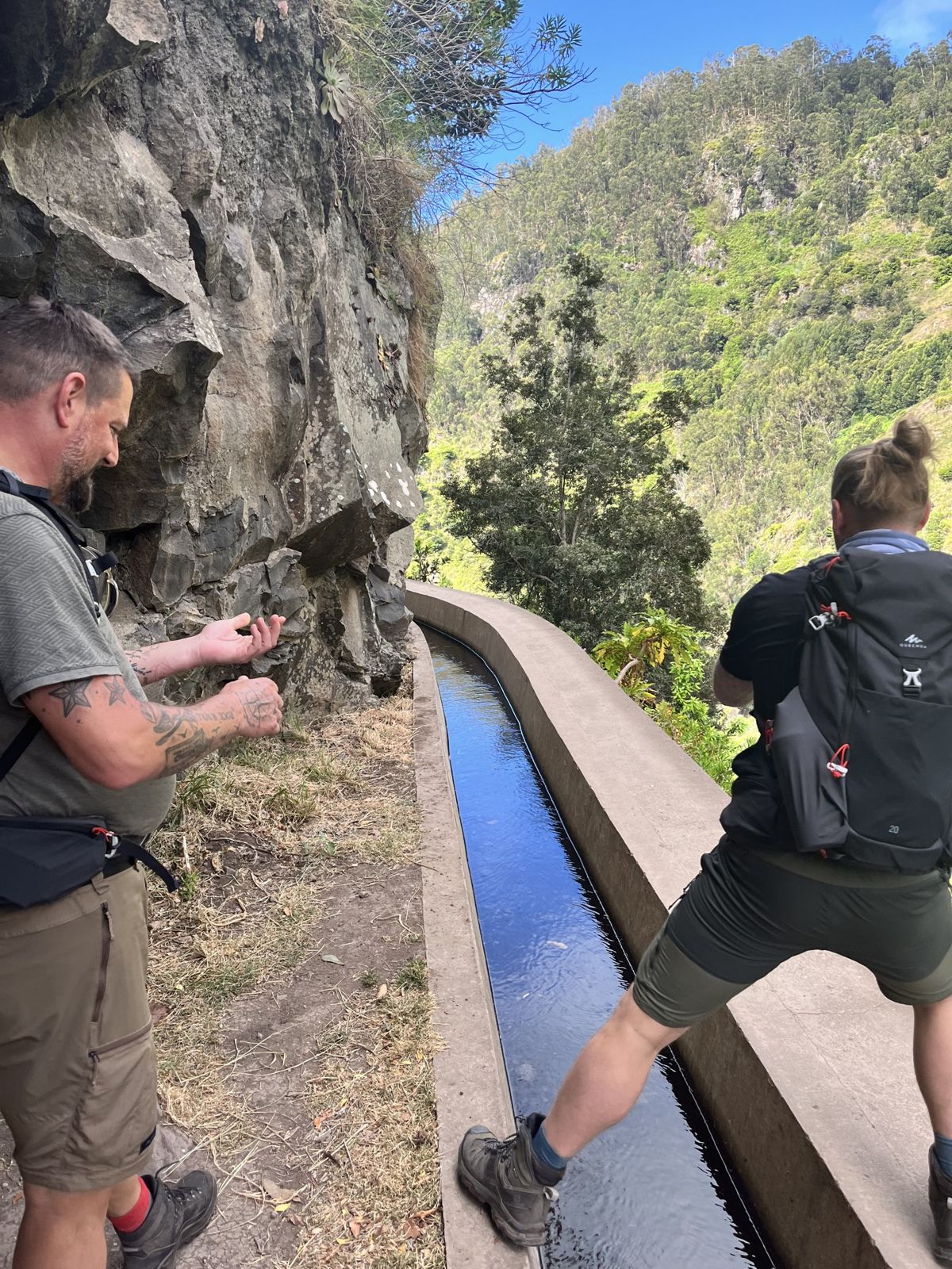 A hiker stepping across a levada water channel while a brother looks on