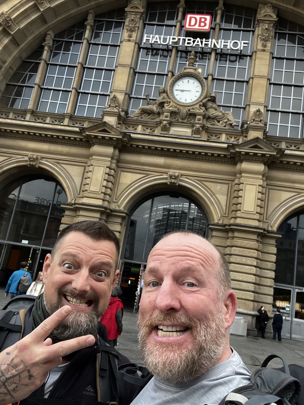 Mike and Nicki grinning in front of the Frankfurt Hauptbahnhof clock and DB sign