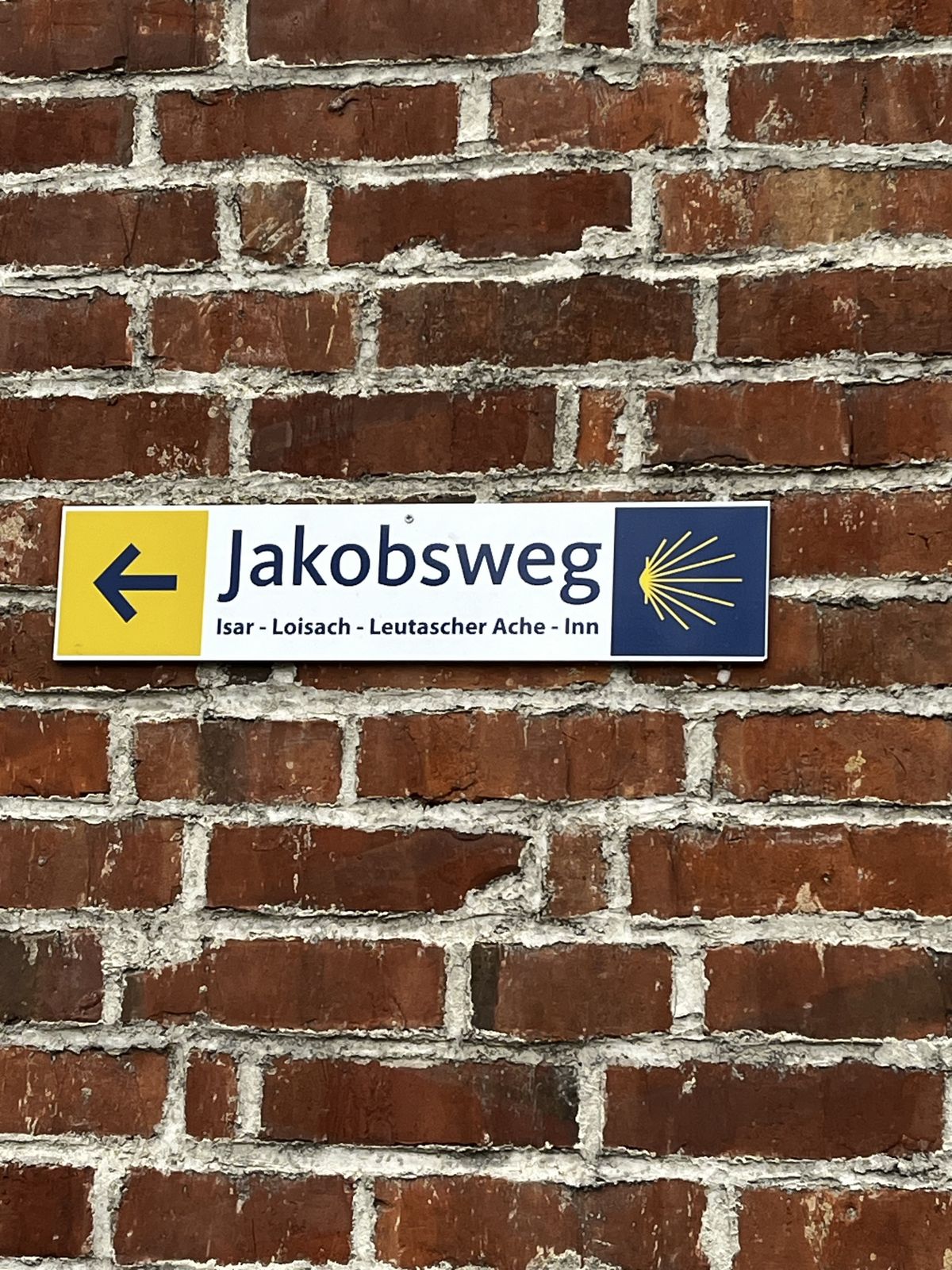 A Jakobsweg waymark sign with yellow arrow and scallop shell on a brick wall