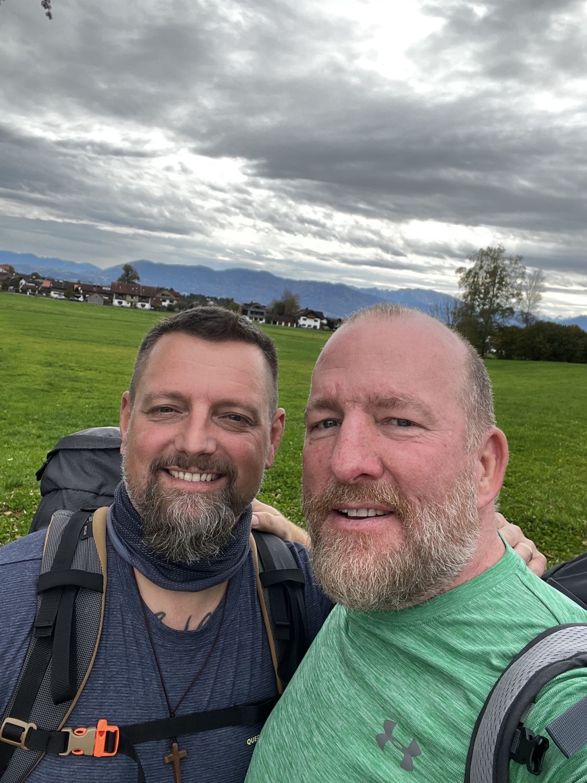 Mike and Nicki selfie in a green meadow with a village and the Alps behind them