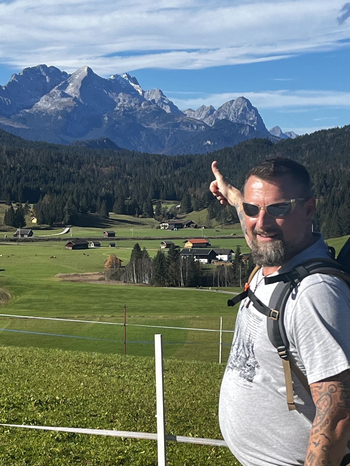 Nicki pointing toward jagged snow-dusted peaks above a green alpine valley