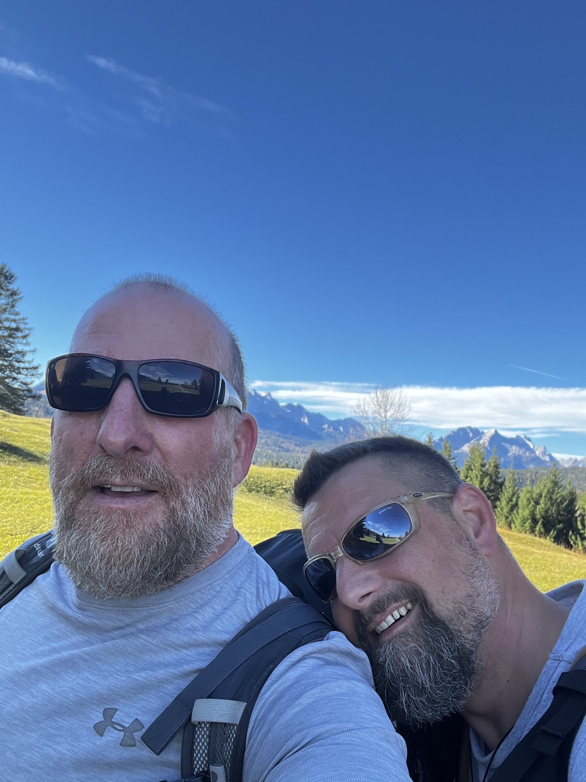 Mike and Nicki in a sunny alpine meadow, Nicki resting his head on Mike's shoulder, peaks behind