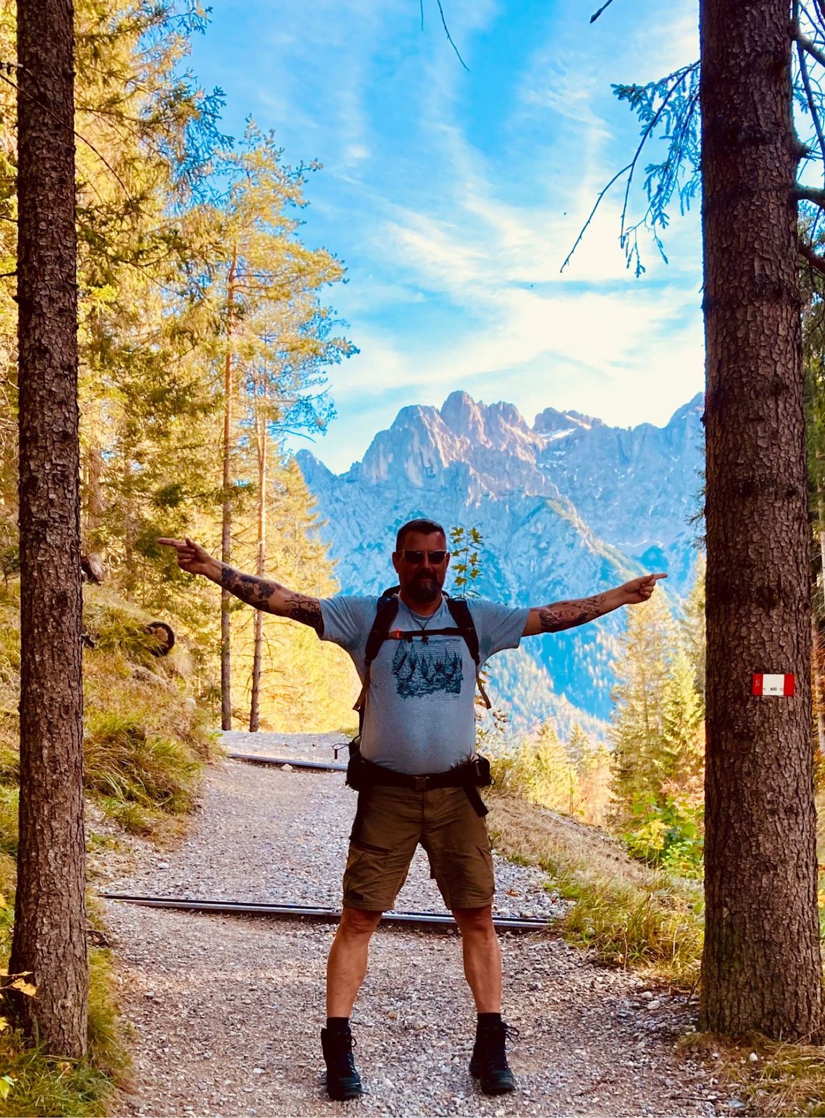 Nicki standing arms outstretched on a forest path with jagged peaks behind