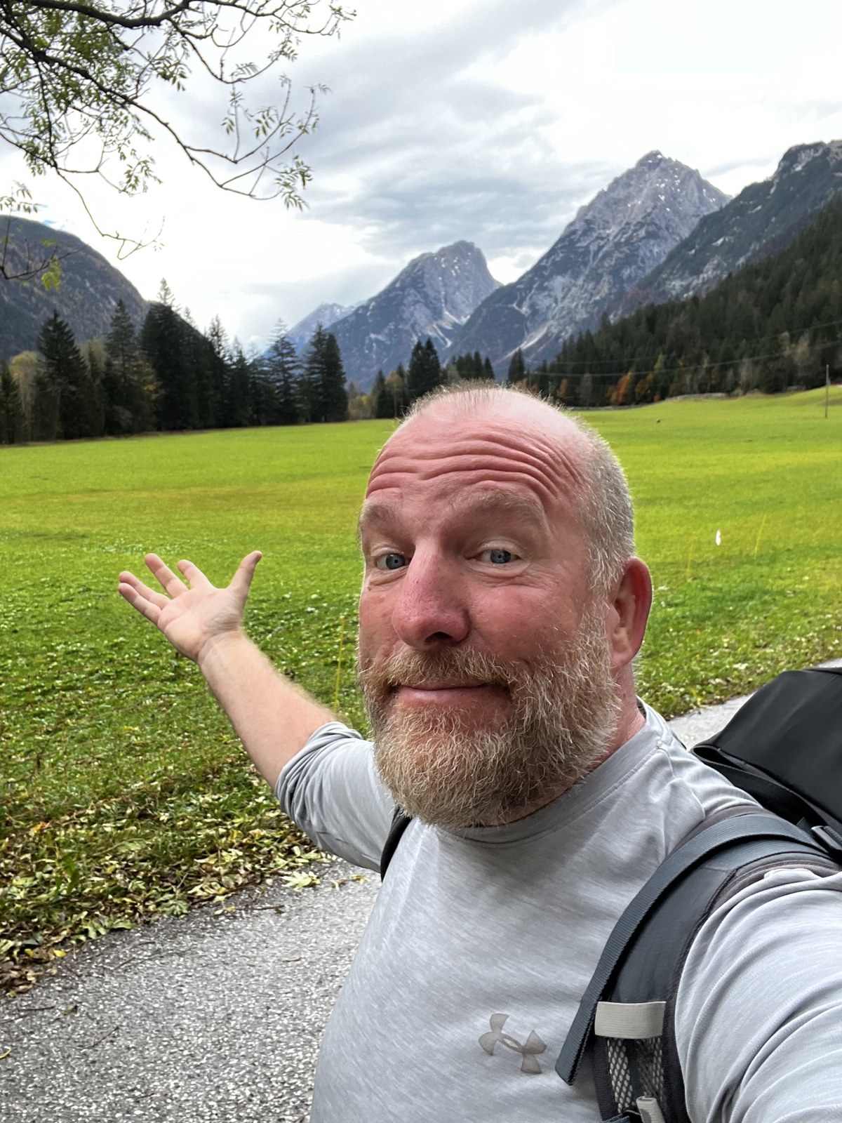 Mike gesturing toward a green alpine meadow with high rocky peaks behind