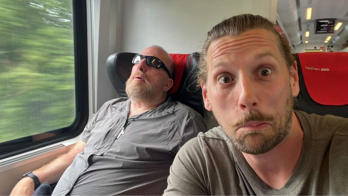 Mike asleep in a train seat wearing sunglasses while Dennis pulls a face in the foreground