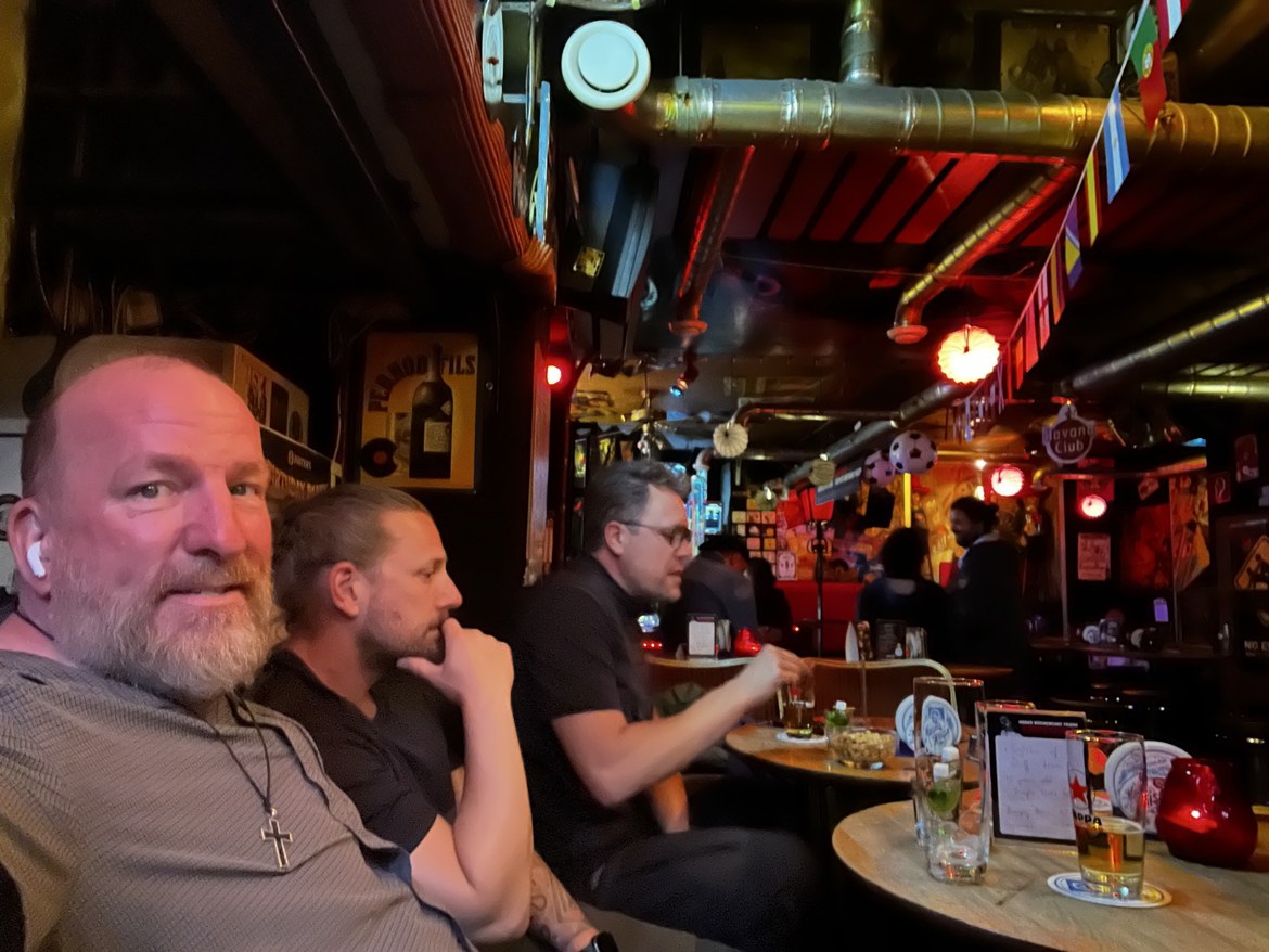 Mike in the foreground of a bar while Dennis and a friend sit at a table behind him, World Cup flags overhead