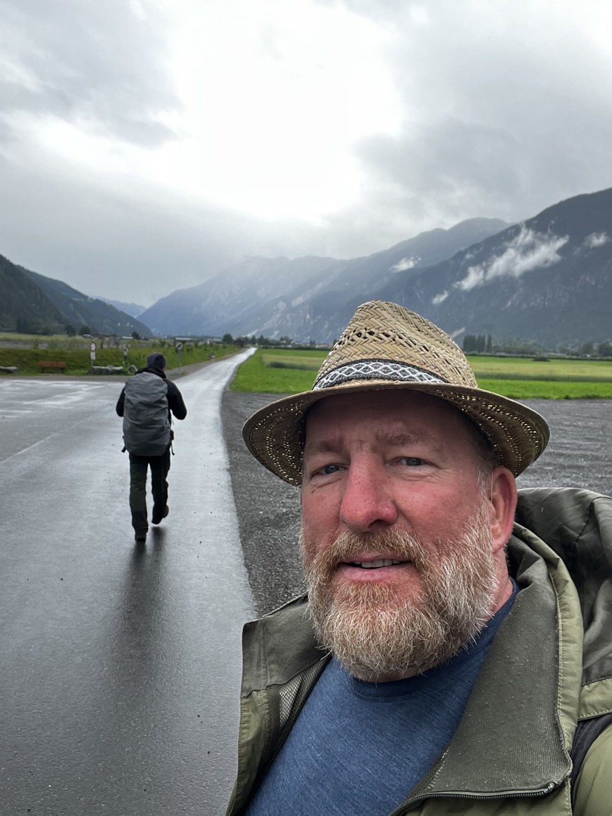 Mike in a straw hat taking a selfie on a wet mountain road, Nicki walking ahead, green valley and peaks behind
