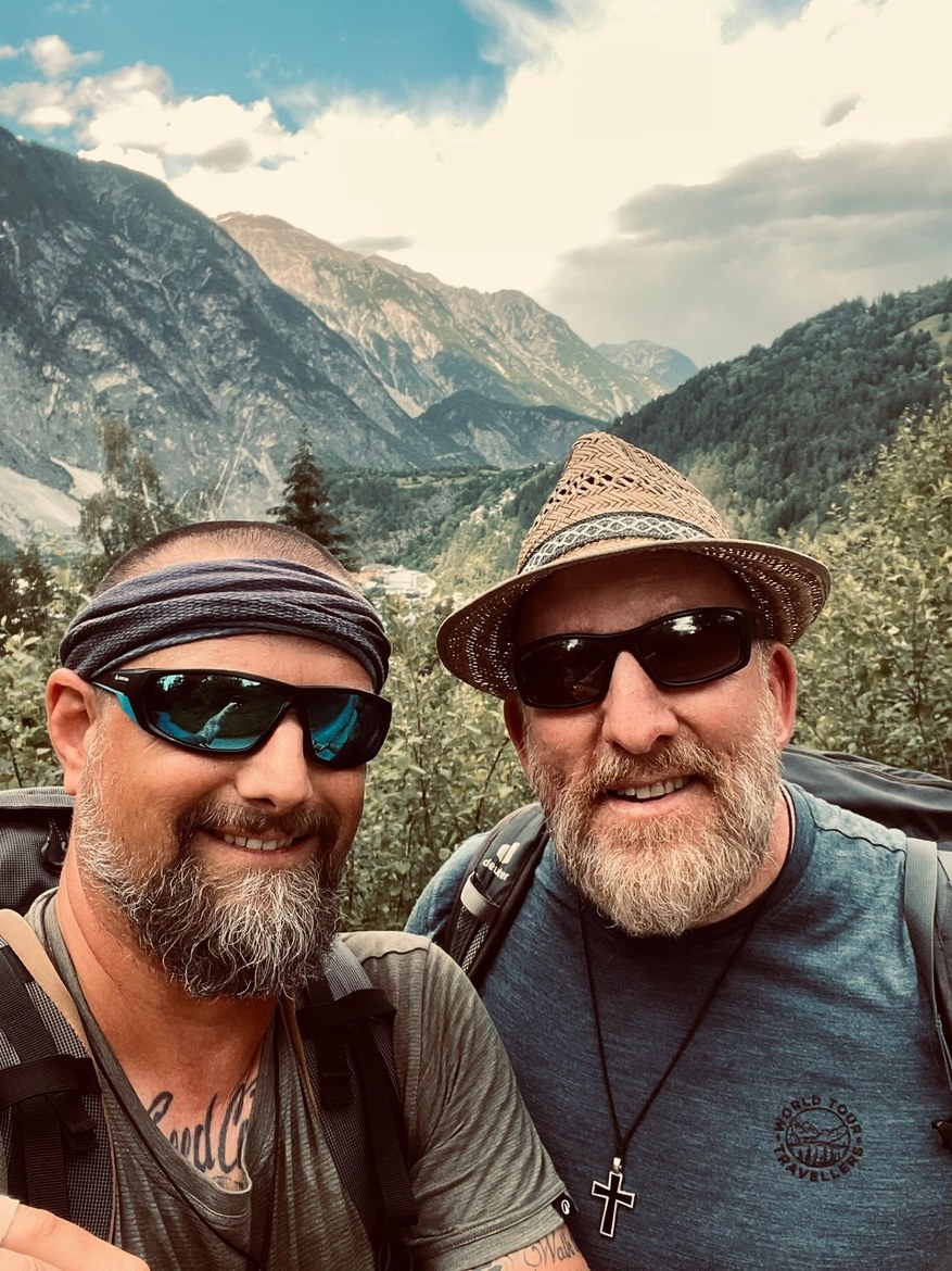 Nicki in a bandana and Mike in a straw hat taking a selfie on the trail, mountains behind