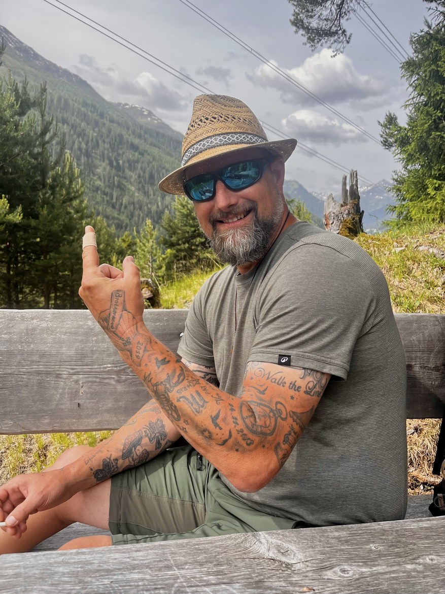 Nicki sitting on a bench in a straw hat and sunglasses, showing a bandaged finger, tattooed arms