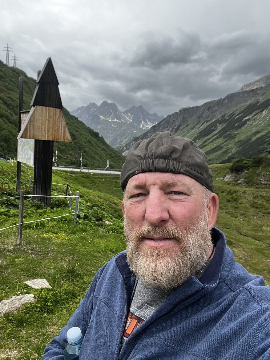 Mike in a cap taking a selfie beside a tall wooden trail signpost, jagged peaks behind